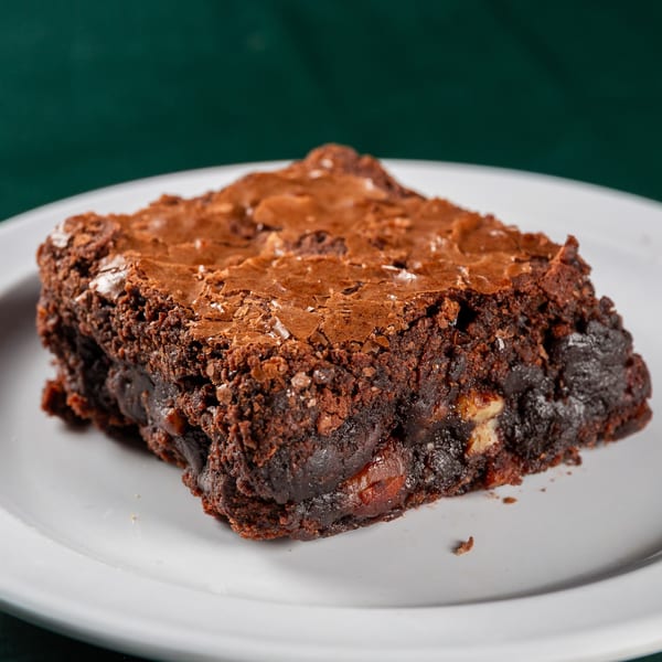 A rich, dark chocolate brownie with a crinkled top and visible chocolate chunks on a white plate.